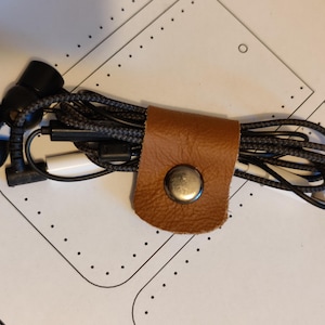 May include: Black earbuds and a braided charging cable are held together by a brown leather cable organizer with a silver snap. The earbuds are on the left, and the cable is neatly coiled.
