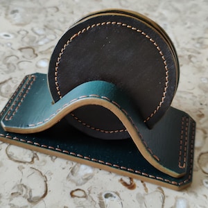 May include: A set of four black leather coasters with a matching holder. The coasters and holder are dark green with tan stitching. The holder has a curved design to hold the coasters.