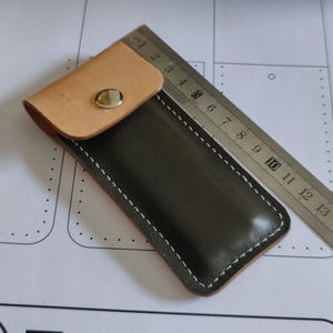 Könnte beinhalten: Eine handgefertigte Ledertasche mit einem naturfarbenen, hellbraunen Überschlag und einem dunkelgrünen Körper. Die Tasche wird mit einem silbernen Druckknopf und weißen Nähten verschlossen. Ein Metalllineal liegt daneben.