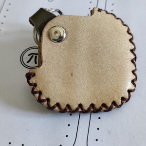 Könnte beinhalten: Ein hellbrauner Leder-Schlüsselanhänger mit dunkelbraunem, genähtem Rand. Der Schlüsselanhänger hat einen silbernen Metallring und eine dunkelgrüne Lederschlaufe. Ein silberner Druckknopf verschließt den Halter.