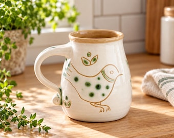 May include: A cream-colored ceramic mug with a rounded body and a sturdy handle. The exterior features a hand-painted brown outline of a bird with green polka dots on its body, accompanied by small green leaf motifs and a tan rim.