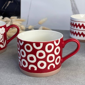Può includere: Tazza in ceramica rossa con motivi circolari bianchi. La tazza ha un manico rosso e una base marrone chiaro. Sono visibili anche altre tazze con disegni diversi. Adatto per bevande calde.
