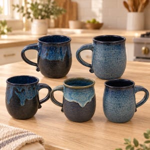 Könnte beinhalten: Ein Set aus fünf Keramikbechern in verschiedenen Blau- und Schwarztönen. Die Becher haben abgerundete Körper, geschwungene Henkel und unterschiedliche Oberflächen, darunter glatte, gesprenkelte und gerippte Texturen mit Tropfglasur.