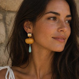 May include: Gold-coloured earrings featuring a rectangular top, a rounded, light-brown centre, and a teardrop-shaped, light-blue bottom. The earrings are worn on a person's ear, with the person's face and hair visible.