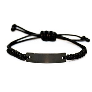 May include: Black adjustable bracelet featuring a rectangular, dark metal plate. The bracelet is made of a woven, black cord with a sliding knot closure for a customizable fit. The plate is centered on the wrist.