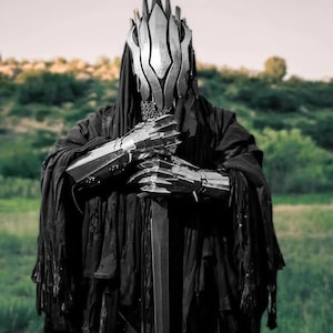 May include: A figure in a black cloak and silver helmet, with matching gauntlets, stands in a field. The helmet has a crown-like design. The figure is holding a long, dark sword. The background is a blurred green field and sky.