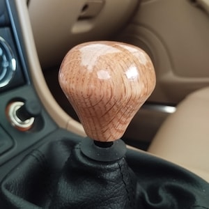 May include: A wooden gear shift knob with a glossy finish. The knob is light brown with visible wood grain and sits atop a black leather shift boot. The interior of the car has tan and black accents.