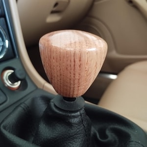 Peut inclure: Pommeau de levier de vitesse en bois clair, monté sur un soufflet de levier de vitesse en cuir noir. L'intérieur de la voiture est beige. Le pommeau est poli.