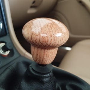 Puede incluir: Pomo de palanca de cambios de madera con forma de hongo. La madera marrón claro tiene un acabado brillante. Está fijado a un fuelle de palanca de cambios de cuero negro. El interior del coche tiene detalles en beige y beige.