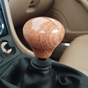 Puede incluir: Un pomo de palanca de cambios de madera con acabado brillante. El pomo es de color marrón claro con veta de madera visible y se asienta sobre una funda de palanca de cambios de cuero negro. El interior del coche tiene detalles en color beige.