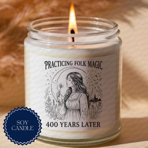 May include: A clear glass jar soy candle with a lit wick. The label features a black and white illustration of a person holding a dandelion, with the text "PRACTICING FOLK MAGIC" at the top and "400 YEARS LATER" at the bottom, plus a dark blue seal.