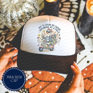 May include: A white and black trucker cap featuring a graphic of a duck wearing sunglasses and a bow, riding a scooter. The duck holds a drink and is surrounded by softball gear. The text reads, OFF TO SPEND MY ENTIRE SATURDAY AT A FIELD.