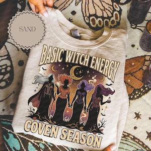 May include: A sand-colored crewneck sweatshirt featuring a graphic print with the text "BASIC WITCH ENERGY" and "COVEN SEASON." The illustration depicts four silhouetted figures in long dresses against a starry night sky with a crescent moon.
