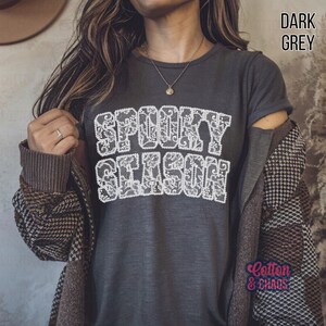 May include: Dark grey t-shirt with the words "SPOOKY SEASON" in a white, lace-like font. The shirt is part of a fall or Halloween-themed collection. The words "Cotton & Chaos" are also visible.