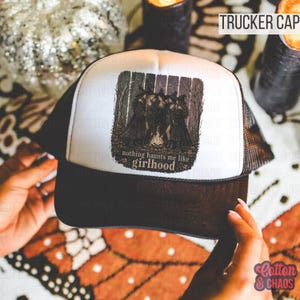May include: A black and white trucker cap with a mesh back. The front panel features a graphic of witches around a fire with the text "nothing haunts me like girlhood". The cap is held by hands.