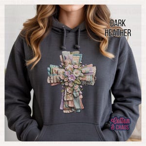 May include: Dark heather gray hoodie featuring a cross design made of layered fabric and flowers. The cross is decorated with pastel colors and floral accents. The text "DARK HEATHER" is printed on the upper right side of the hoodie.