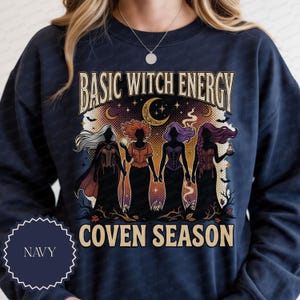 May include: A navy blue sweatshirt featuring a graphic print with the text "BASIC WITCH ENERGY" and "COVEN SEASON". The design displays four silhouetted figures with long hair, wearing corsets and capes, set against a background of stars, a crescent moon, and bats.