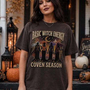 May include: A dark charcoal gray short-sleeve t-shirt featuring a graphic print of four silhouettes of figures in long dresses. The text reads BASIC WITCH ENERGY at the top and COVEN SEASON at the bottom, with a crescent moon and stars above.