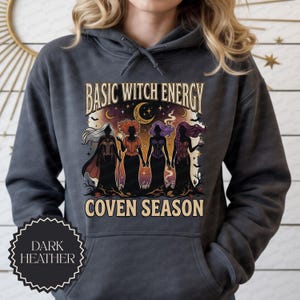 May include: A dark heather gray hooded sweatshirt featuring a graphic print of four silhouetted figures with long hair, a crescent moon, and bats. The text reads BASIC WITCH ENERGY and COVEN SEASON in a decorative font above and below the figures.