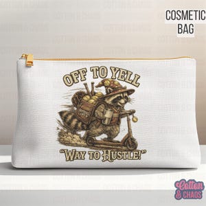 May include: A white cosmetic bag featuring a graphic of a raccoon wearing a hat, carrying a bag of softballs, and riding a scooter. The text reads OFF TO YELL and WAY TO HUSTLE in a brown font. The bag has a gold zipper pull on the top right side.