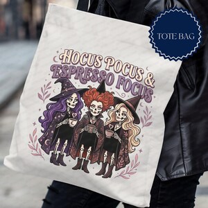 May include: A white tote bag featuring a graphic illustration of three skeleton witches with purple, red, and blonde hair, each holding a coffee cup. The text HOCUS POCUS & ESPRESSO FOCUS is printed in a purple arched font above the figures.