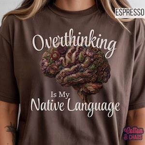 May include: Espresso-colored t-shirt with the text "Overthinking Is My Native Language" in white. The graphic features a detailed illustration of a brain composed of flowers in shades of purple, brown, and green. The shirt is from Cotton & Chaos.