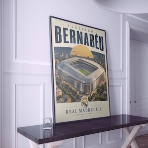 Puede incluir: Un póster vertical enmarcado que presenta una ilustración aérea del estadio Santiago Bernabéu. El texto dice SANTIAGO BERNABÉU en la parte superior y REAL MADRID C.F. MADRID ESPAÑA EST. 1947 en la inferior, con el escudo centrado debajo.