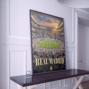 Puede incluir: Un póster vertical enmarcado que muestra una vista aérea de un estadio de fútbol lleno durante el atardecer. La imagen presenta un campo verde, gradas repletas y el texto REAL MADRID en letras doradas grandes, con el escudo del club debajo.