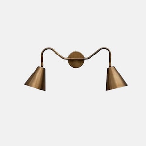 May include: A brass-toned wall sconce with two conical shades. The fixture has a circular base and a curved arm design, with each shade pointing outwards. The sconce is made of metal and has a vintage aesthetic.
