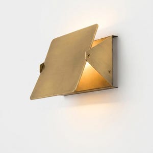 May include: A wall-mounted light fixture crafted from brushed brass metal. The design features a flat, rectangular front plate angled outward, with side panels secured by visible screws, emitting a warm, soft glow from the interior space.
