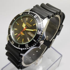 Può includere: Un orologio da polso automatico Citizen con quadrante nero, lunetta girevole nera e cinturino in gomma nero. Il quadrante mostra il giorno e la data, con lancette e indici dorati. L'orologio è esposto su un supporto trasparente.