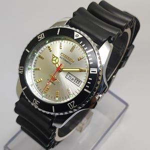 Op de afbeelding: Een Citizen Automatic horloge met een zilveren wijzerplaat, zwarte lunette en zwarte rubberen band. De wijzerplaat toont de dag en datum, met goudkleurige wijzers en markeringen. De lunette heeft witte cijfers. Het horloge wordt op een transparante standaard getoond.
