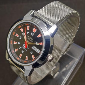 May include: A Citizen automatic wristwatch with a black dial, orange accents, and a silver mesh band. The watch face displays the day and date, with the text "21 Jewels" and "Citizen Automatic". The watch has a stainless steel case.