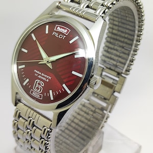 Op de afbeelding: Een zilverkleurig polshorloge met een bordeauxrode wijzerplaat. De wijzerplaat heeft witte markeringen en wijzers, met de tekst "hmk PILOT" en "PARA SHOCK 17 JEWELS INDIA". Het horloge heeft een zilveren, rekbare band.