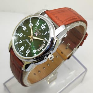 Könnte beinhalten: Eine silberfarbene Seiko Automatik-Uhr mit grünem Zifferblatt und braunem Lederarmband. Das Zifferblatt zeigt weiße Ziffern und Zeiger mit dem Seiko-Logo. Das Armband ist rotbraun.