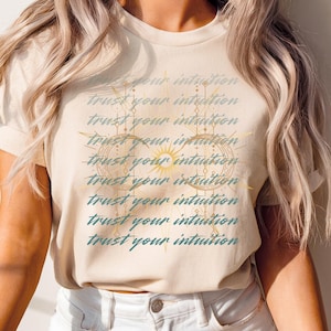 May include: Cream-colored t-shirt with the text "trust your intuition" repeated in a pattern. The design includes a sun and geometric shapes in gold and teal. The shirt is made of a soft material.