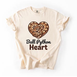 Könnte beinhalten: Ein cremefarbenes Kurzarm-T-Shirt mit einer Grafik eines braun-beigen Königspythons, der sich zu einer Herzform zusammenrollt. Unter der Schlangenillustration ist der Text "Ball Python Heart" in einer dunkelbraunen, fetten Schrift gedruckt.