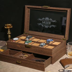 May include: A wooden watch box with a glass top, displaying several wristwatches. The box has two levels, with a drawer at the bottom. The top of the box has the word "BELEK" in a decorative font. The box is brown with a gold clasp.
