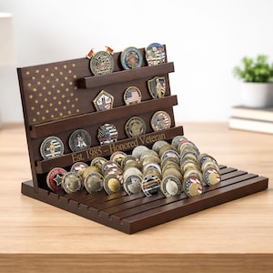 Op de afbeelding: Een houten muntendisplay met meerdere planken, met talrijke herdenkingsmunten. De standaard heeft een Amerikaans vlagontwerp en de tekst "Est. 1985 - Honored Veteran". De munten hebben verschillende ontwerpen en kleuren.