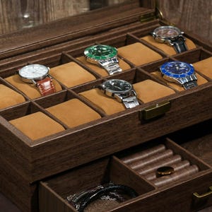 May include: A wooden watch box with a clear lid, showcasing several wristwatches with various colored faces and bands. The box has multiple compartments with tan cushions, and a lower drawer for additional storage.