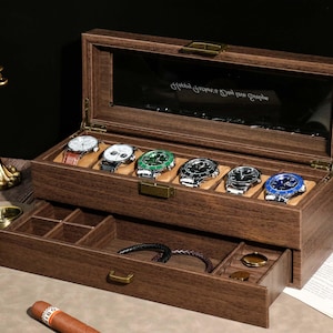 May include: A wooden watch box with a glass top, displaying six wristwatches with various colored faces and straps. The box has a lower drawer for additional storage. The watches are displayed on a tan cushion.