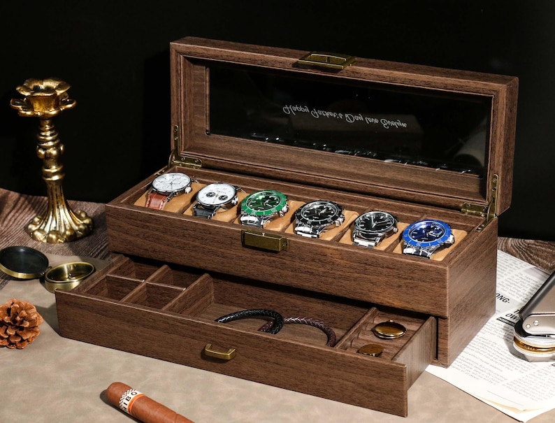 May include: A wooden watch box with a glass top, displaying six wristwatches with various colored faces and straps. The box has a lower drawer for additional storage. The watches are displayed on a tan cushion.