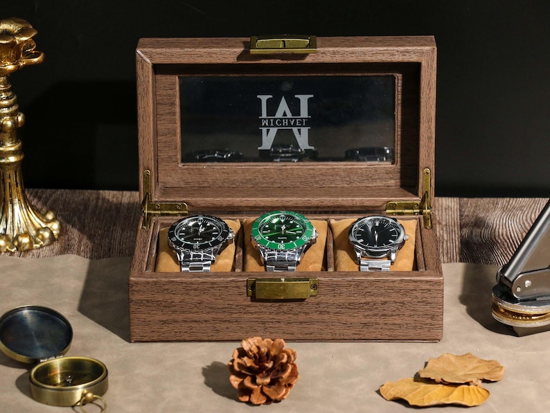 May include: A wooden watch box with three watches inside. The watches have silver bands and black or green faces. The box has a glass top with the name "WICHVET" printed on it. The box is open, revealing the watches.