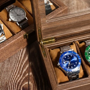 May include: Wooden watch display case with multiple compartments, each holding a silver wristwatch. Two watches have black faces, while two others have blue and green faces. The case is open, revealing the watches.