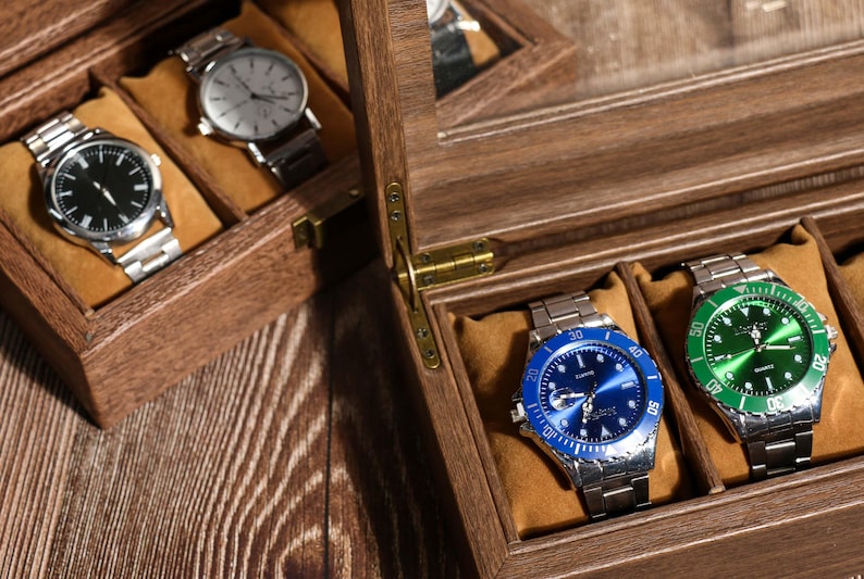 May include: Wooden watch display case with multiple compartments, each holding a silver wristwatch. Two watches have black faces, while two others have blue and green faces. The case is open, revealing the watches.