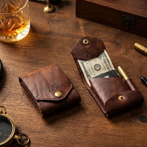May include: Two dark brown leather wallets with a distressed finish and brass snap closures. One wallet is closed and features the embossed initials J.K., while the other is open, revealing a paper dollar bill, a black card, and a brass pen.