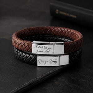 以下が含まれることがあります： 茶色と黒の2つの編み込みレザーブレスレット。シルバーの留め金が付いています。茶色のブレスレットには「Patrick love you forever Dad」と刻まれています。黒いブレスレットには「I love you Daddy」と書かれています。