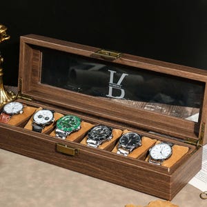 May include: A wooden watch display case with a glass top, holding six assorted wristwatches. The case is open, revealing watches with different colored faces and bands. The interior is lined with a soft, tan material. The top of the case has the letters "V DOBELL" etched into the glass.