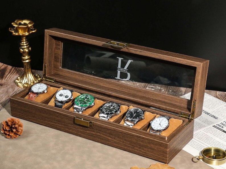 May include: A wooden watch display case with a glass top, holding six assorted wristwatches. The case is open, revealing watches with different colored faces and bands. The interior is lined with a soft, tan material. The top of the case has the letters "V DOBELL" etched into the glass.