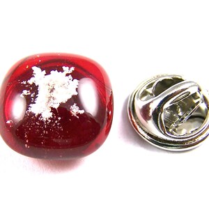 May include: A red glass lapel pin with a silver-colored pin back. The glass has a rounded shape and contains a white, cloud-like design. The pin is set against a white background.